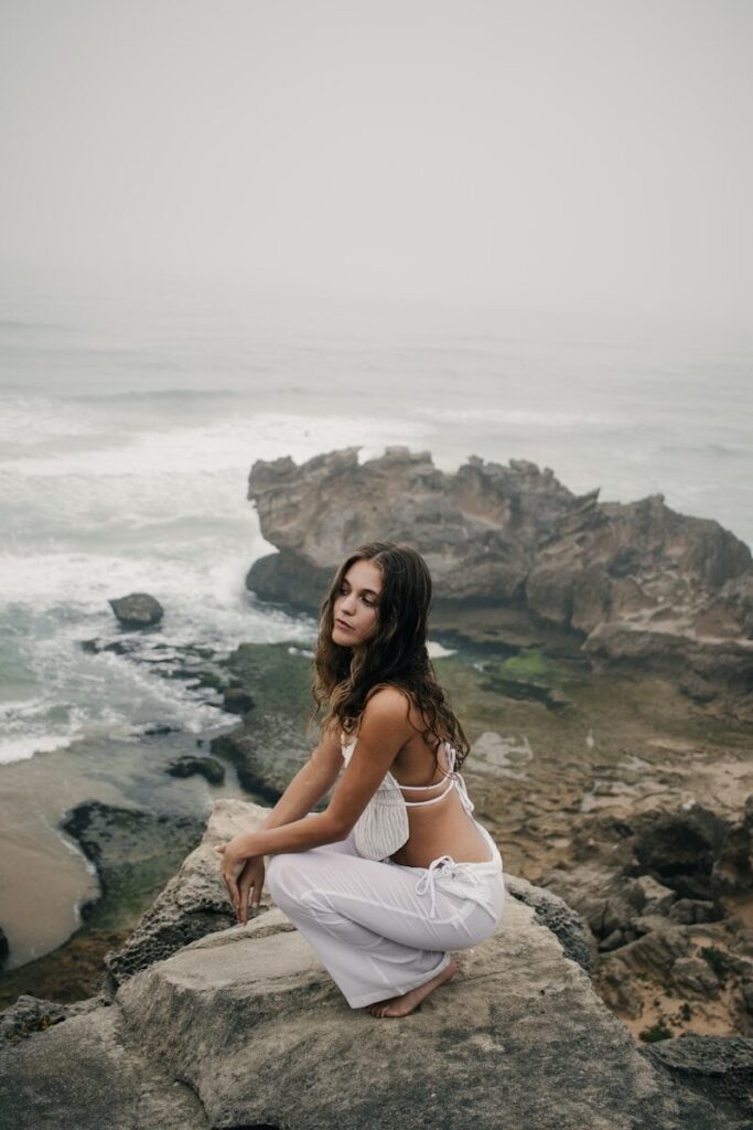 Woman in white clothes crouching on rocks by the ocean