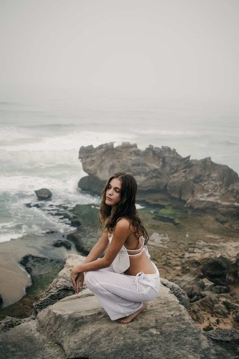 Woman in white clothes crouching on rocks by the ocean